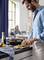 A man is chopping vegetables on a chopping board in a kitchen. A woman is sitting at the table in the background.