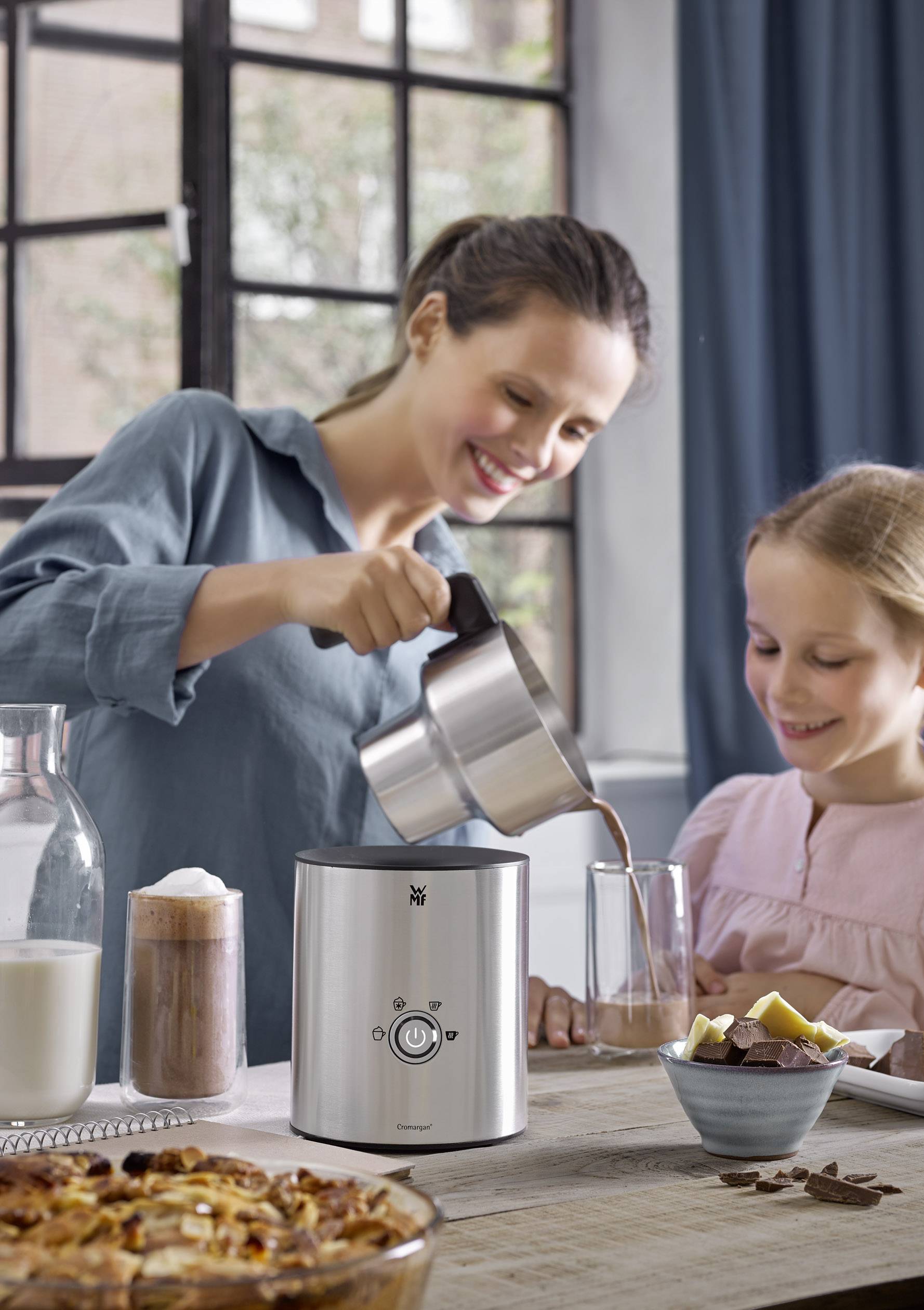 A woman is pouring a hot drink into a glass, while a girl stands next to her and smiles. Milk, drinks, and snacks are on the table.