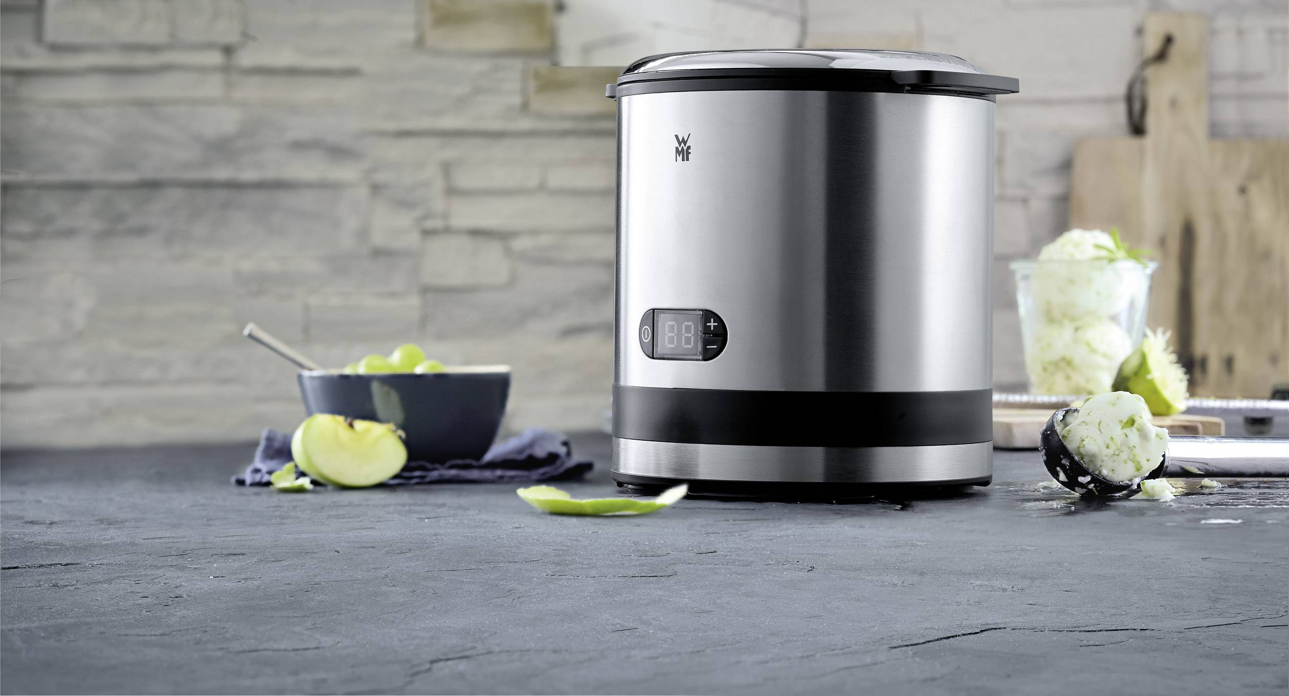 Stainless steel kitchen appliance on a worktop, alongside fruit, a bowl, and a grater. Stone-effect wall in the background.