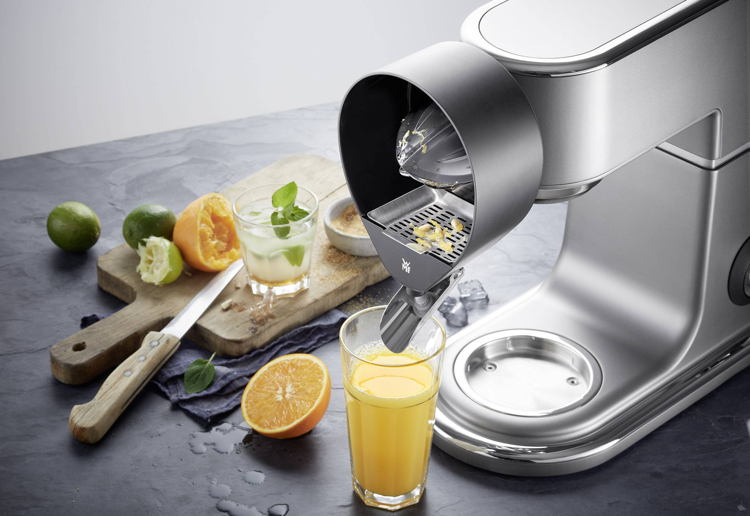 A silver food processor with a juicer presses fresh orange juice into a glass. Citrus fruits are scattered in the background.