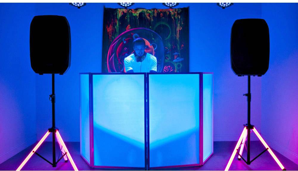 A DJ is playing music in a brightly lit room, flanked by large speakers. The lighting creates a dynamic atmosphere.