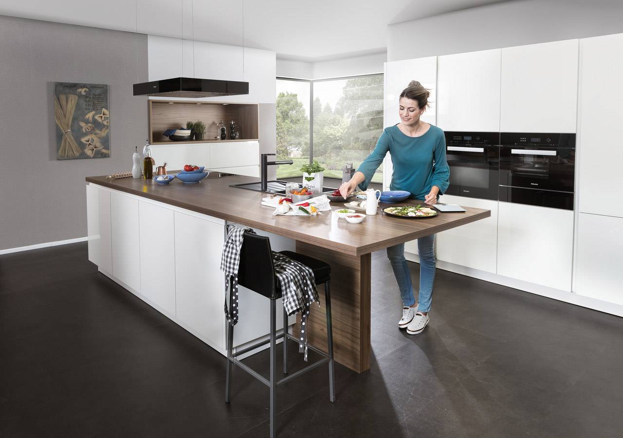 A woman is preparing food in a modern, bright kitchen. Spices and ingredients are on the worktop, with a large window in the background.