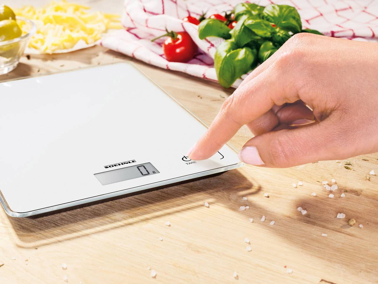 A hand presses the power button of a digital kitchen scales on a wooden table. Tomatoes and spices are visible in the background.
