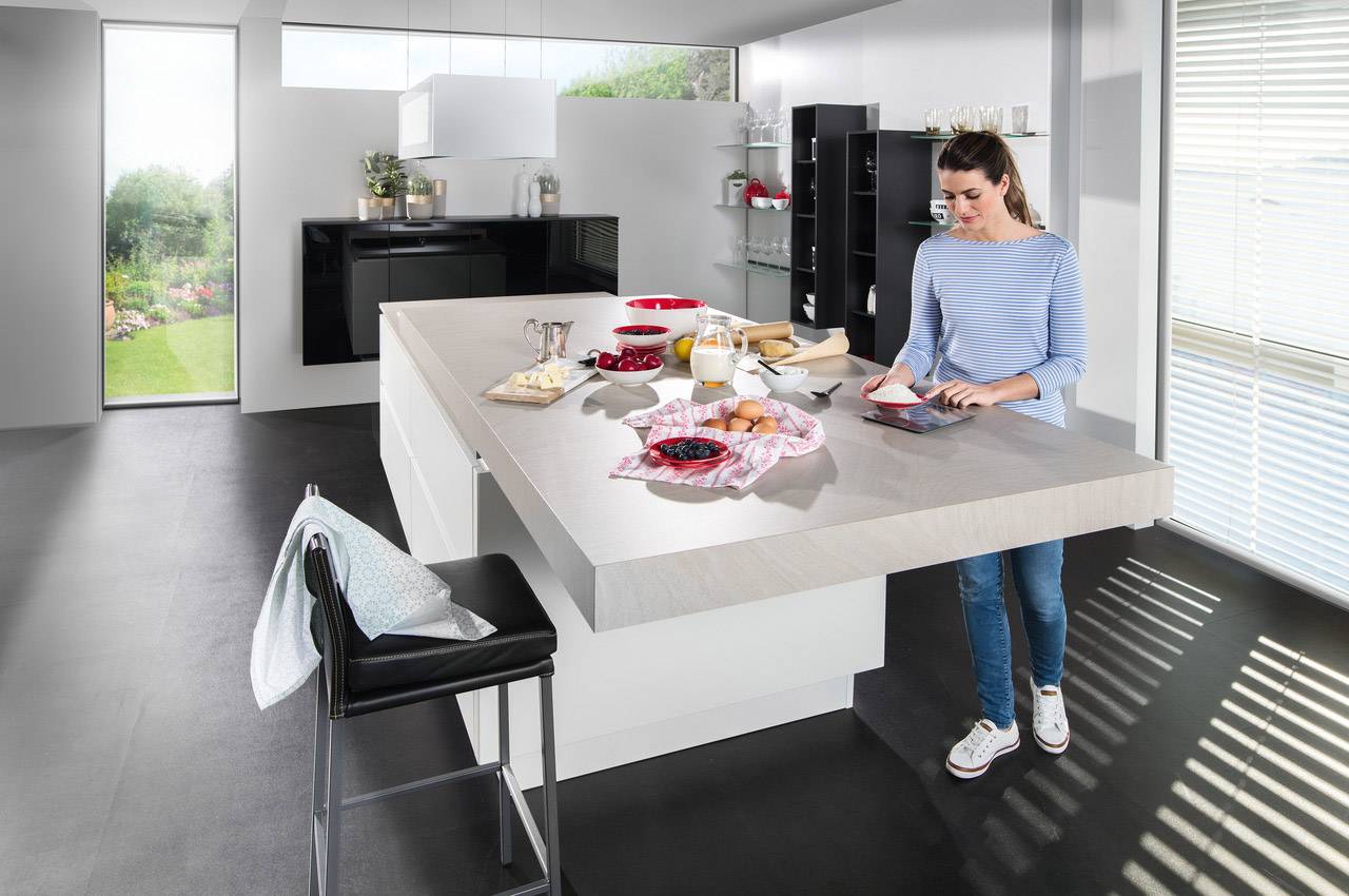 A woman stands in a modern kitchen, preparing a dish on a long, grey island. Fruit and a carafe of juice are laid out on the island.