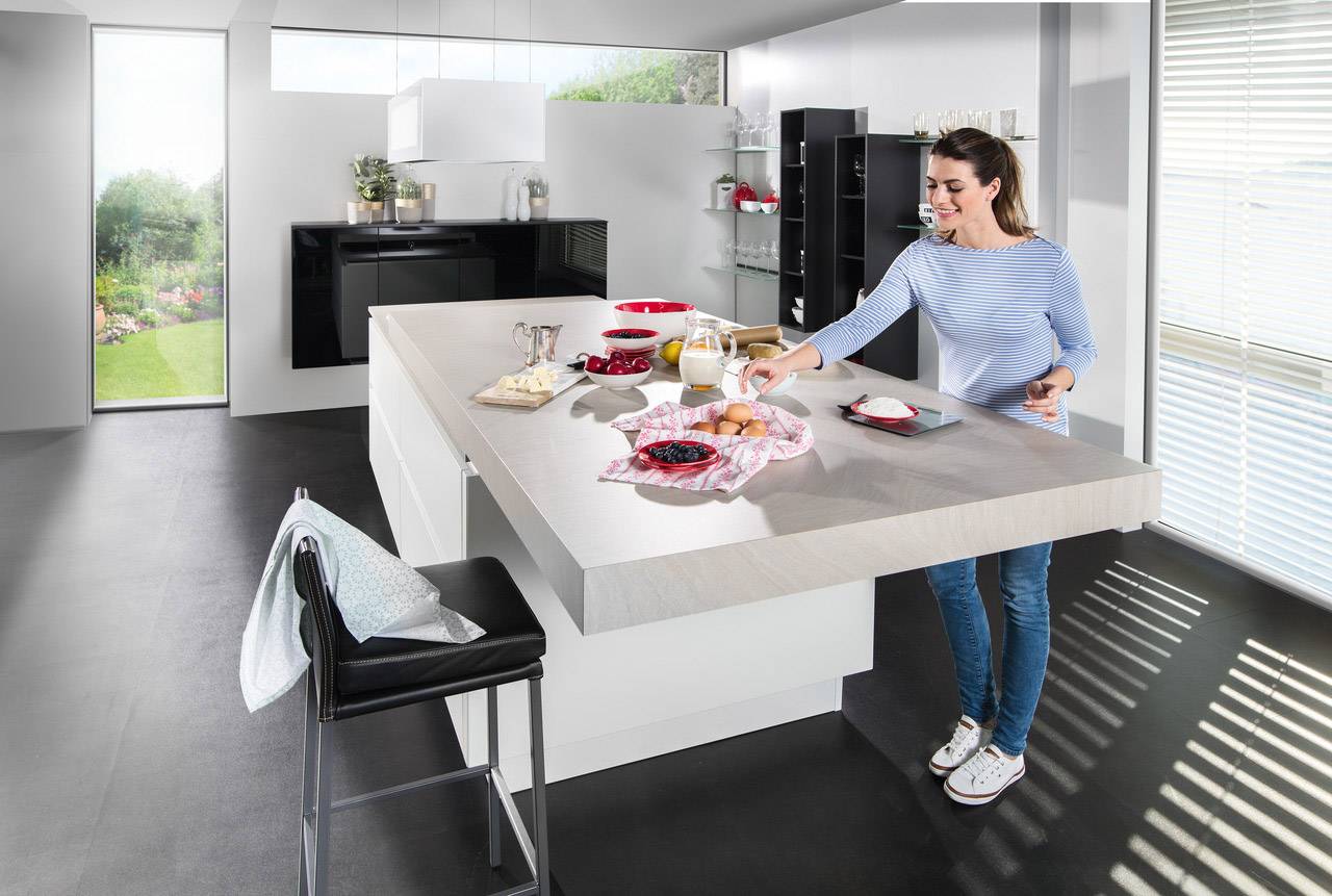 A woman is standing at a modern kitchen worktop, preparing breakfast with toast, jam, orange juice, and eggs.