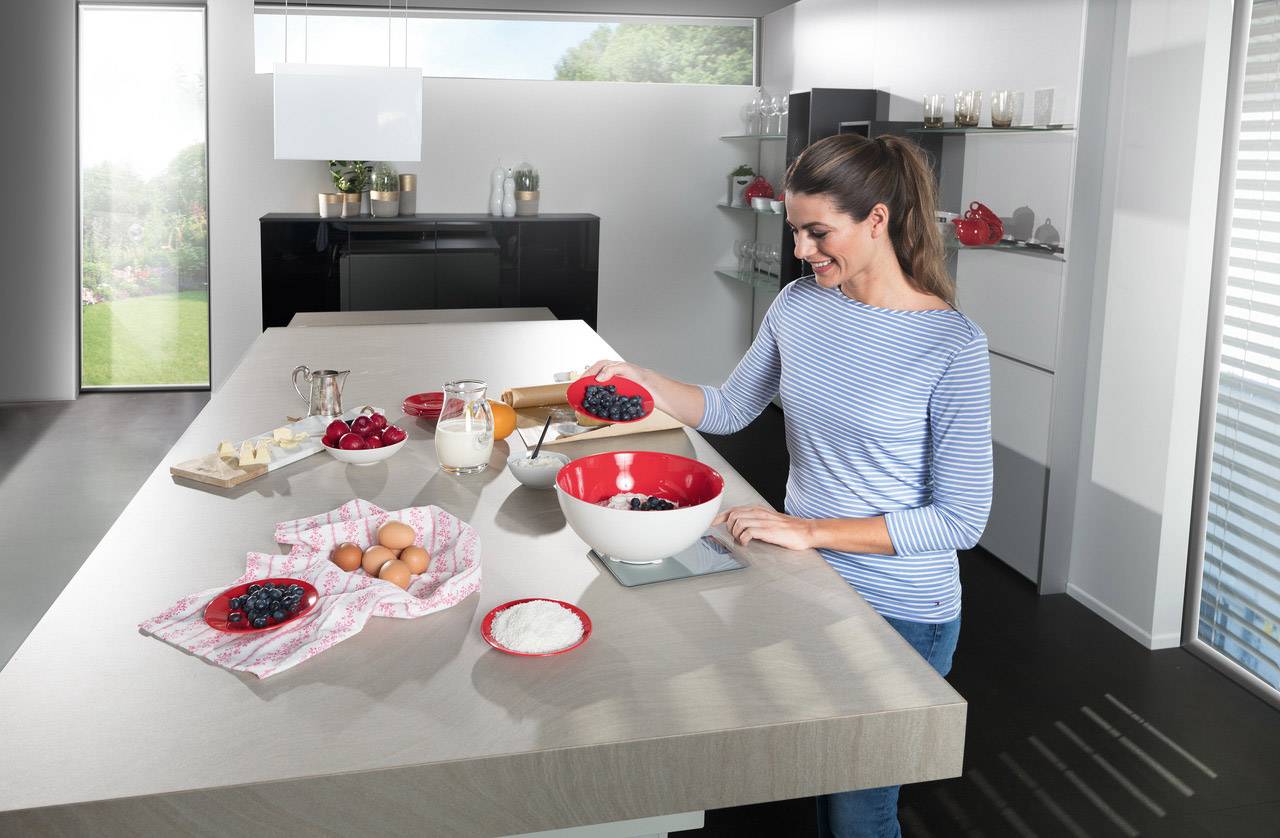A woman in a modern kitchen prepares ingredients such as eggs, milk, and berries for a recipe. She is holding a bowl of blueberries.