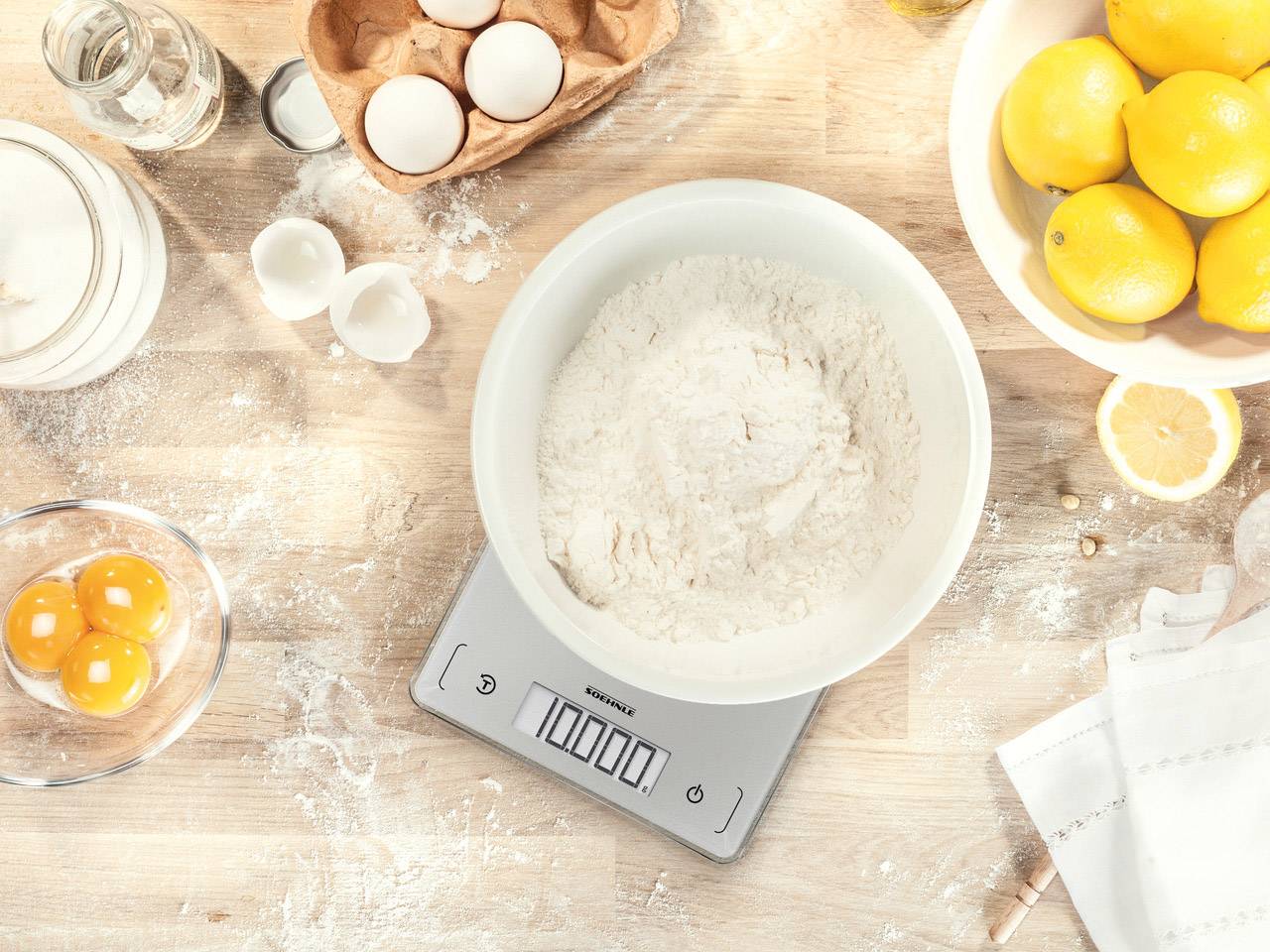 Scales in a kitchen with a bowl of flour showing '1000g', surrounded by eggs and lemons on a wooden board; preparation for baking.