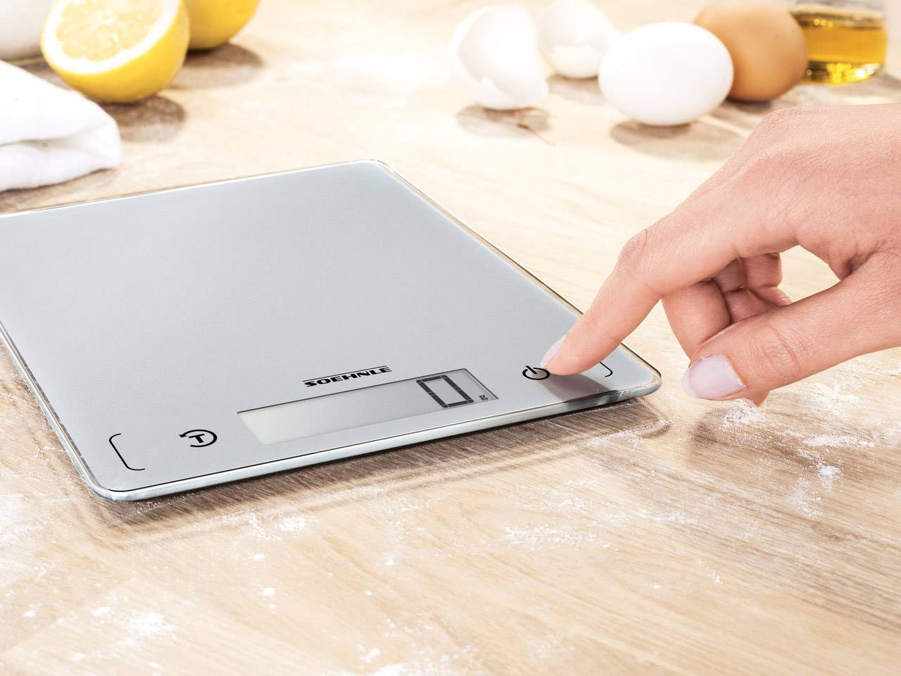 A digital kitchen scale on a table, surrounded by flour, lemons, and eggs. A hand is pressing the power button.