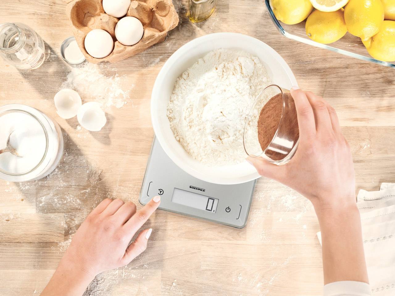 A person is weighing cocoa powder in a bowl with flour on a digital kitchen scale. Eggs and lemons are in the background.