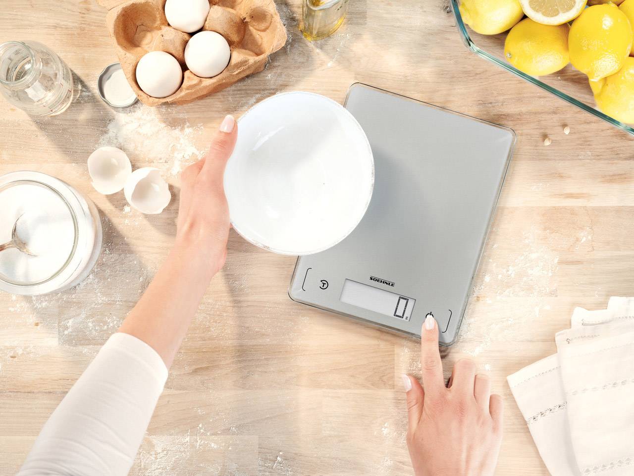 A person is weighing an empty bowl on a digital kitchen scales, surrounded by eggs and lemons on a wooden table.