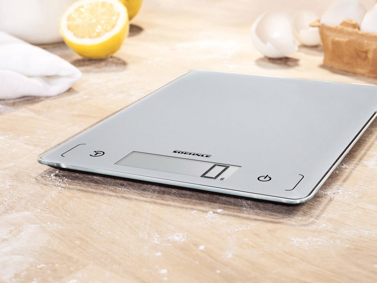 Digital kitchen scales on a flour-dusted worktop with lemons and eggshells in the background.