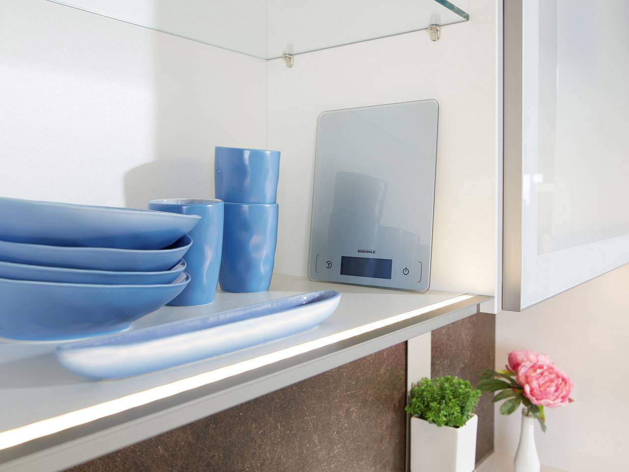 A kitchen scene with a blue crockery collection and a digital kitchen scale. Decoratively placed plants and flowers.