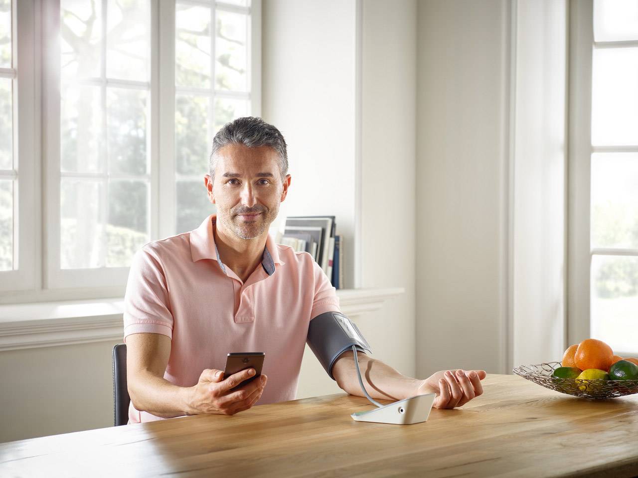 A man in a pink shirt is measuring his blood pressure at home using a digital device. He is holding a smartphone in his hand.