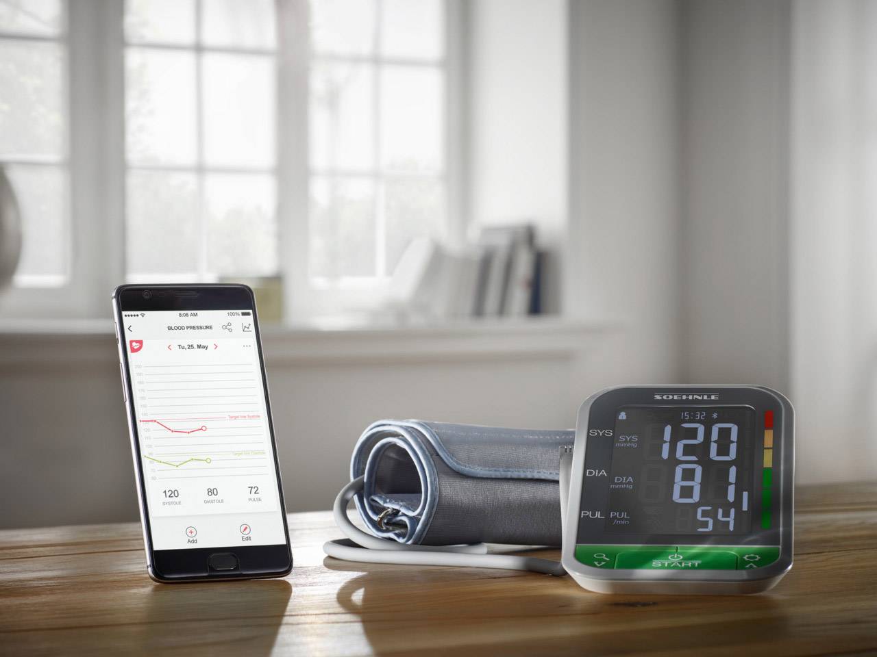 A smartphone and a blood pressure monitor on a table. The smartphone displays a health app with a graph, the device shows a reading of 120/81.