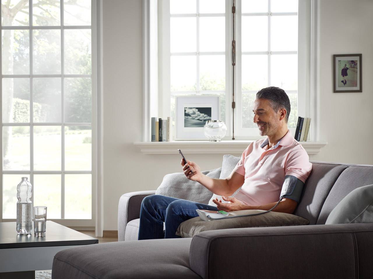 A man is measuring his blood pressure whilst sitting on a sofa. He is looking at a smartphone and wearing a blood pressure cuff on his left arm.