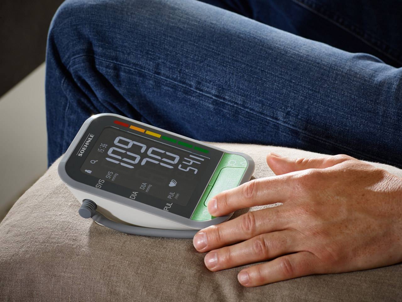 A person is measuring their blood pressure using a digital blood pressure monitor positioned on their thigh. The display is showing the readings.