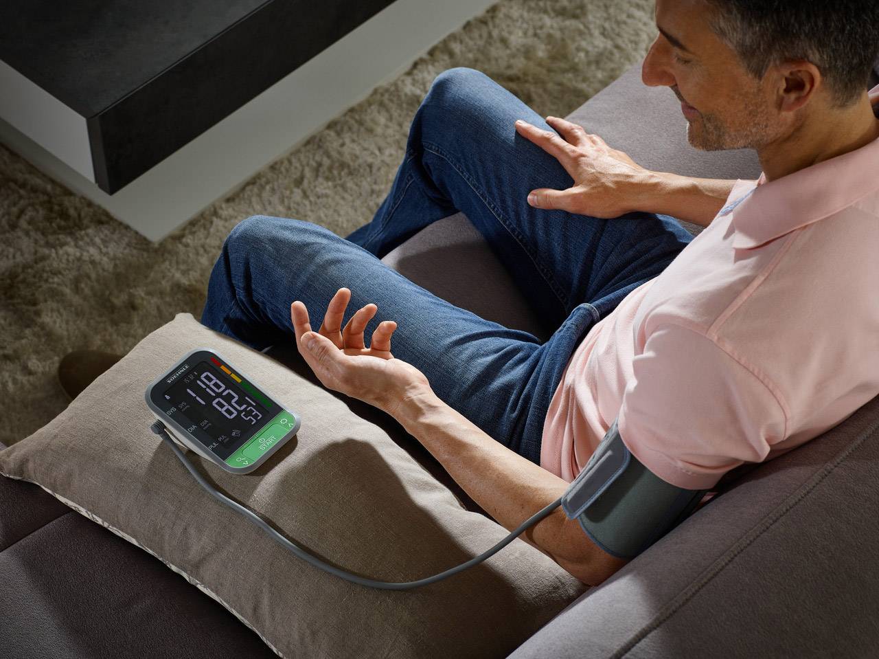A man is sitting on a sofa and measuring his blood pressure with a digital blood pressure monitor attached to his left arm.