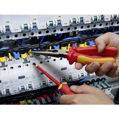 Two hands holding pliers and screwdrivers in a fuse box, surrounded by numerous cables and switches, for maintenance.