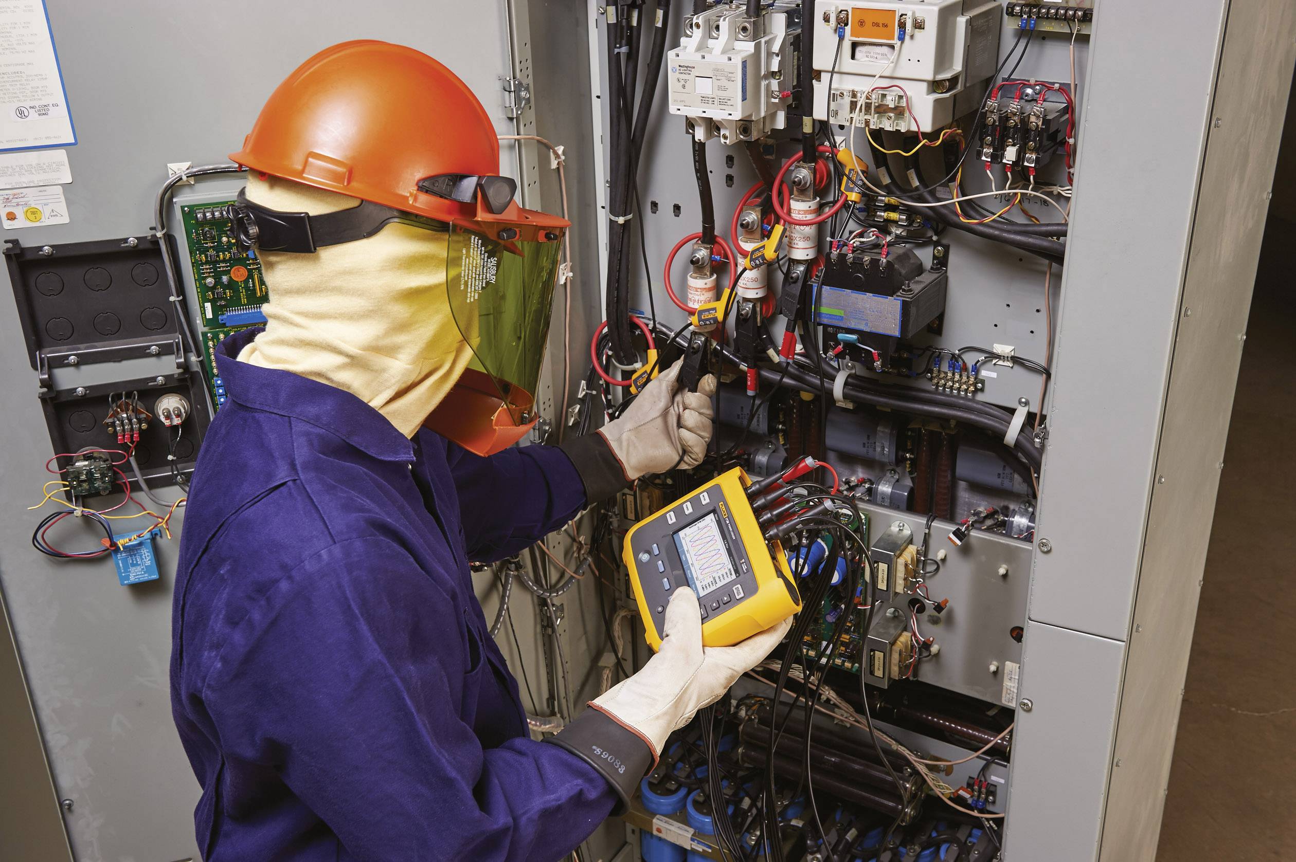 An electrician in protective gear is checking the wiring in an open electrical cabinet with a measuring device.