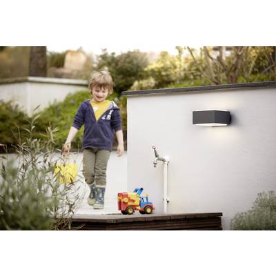 A child with a yellow bucket walks in the garden. In front of them stands a colourful toy car. A wall light shines in the background.