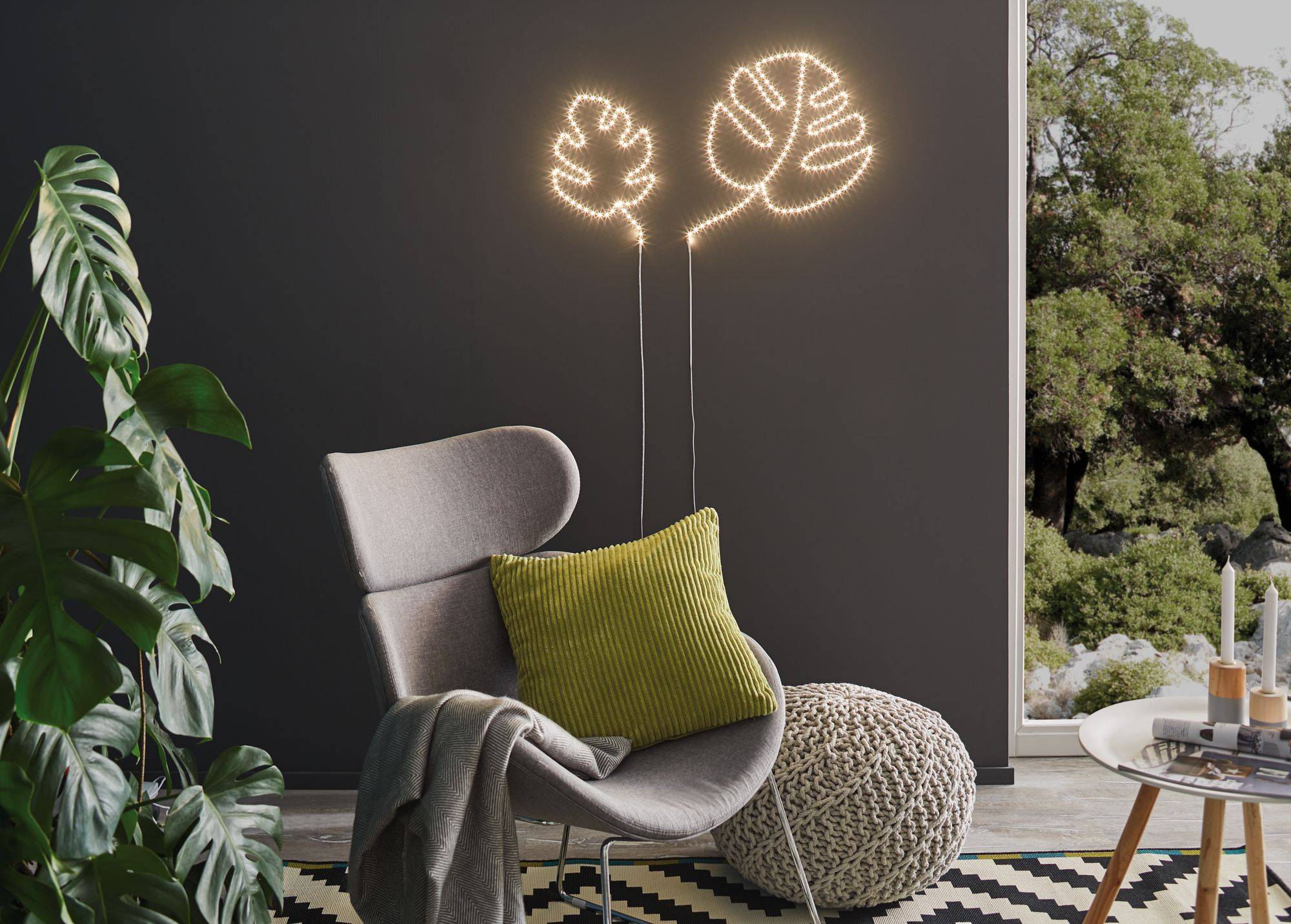 A grey armchair with a green cushion in front of a dark wall with illuminated plant motifs. A Monstera plant stands to the left.
