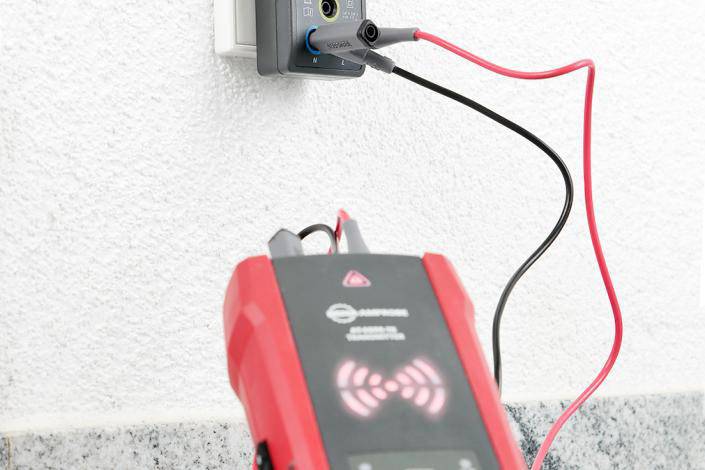 A voltage tester is plugged into a socket. A red device with cables indicates that the electrical circuit is live.