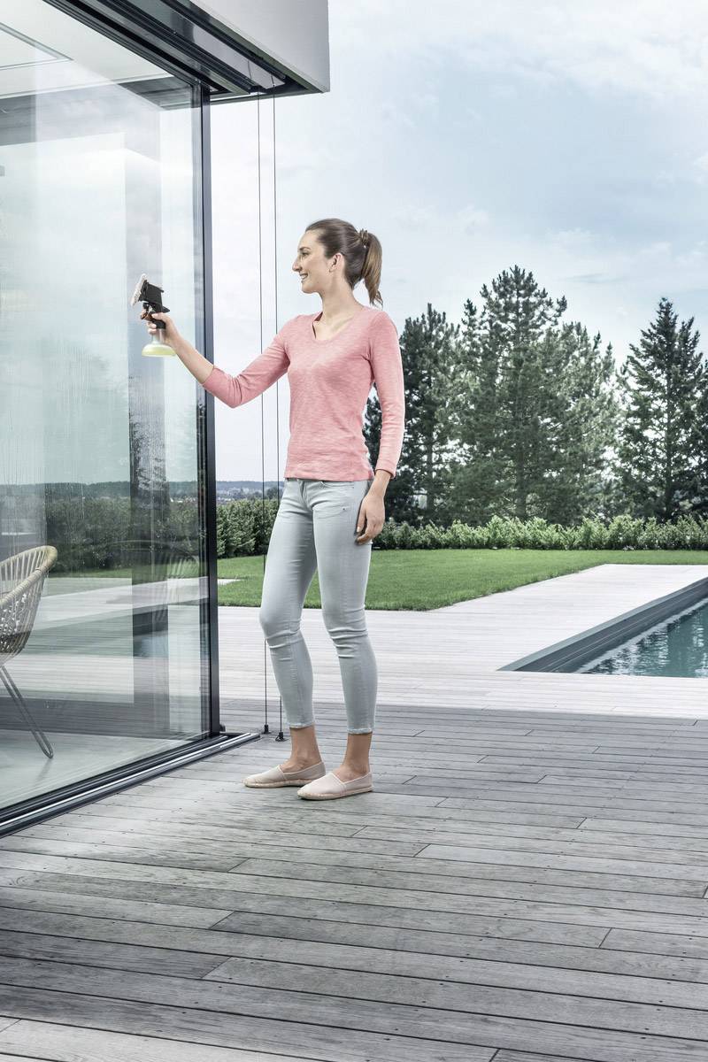 A woman is cleaning glass with a spray and cloth on a terrace with a swimming pool in the background. Trees and sky are visible.