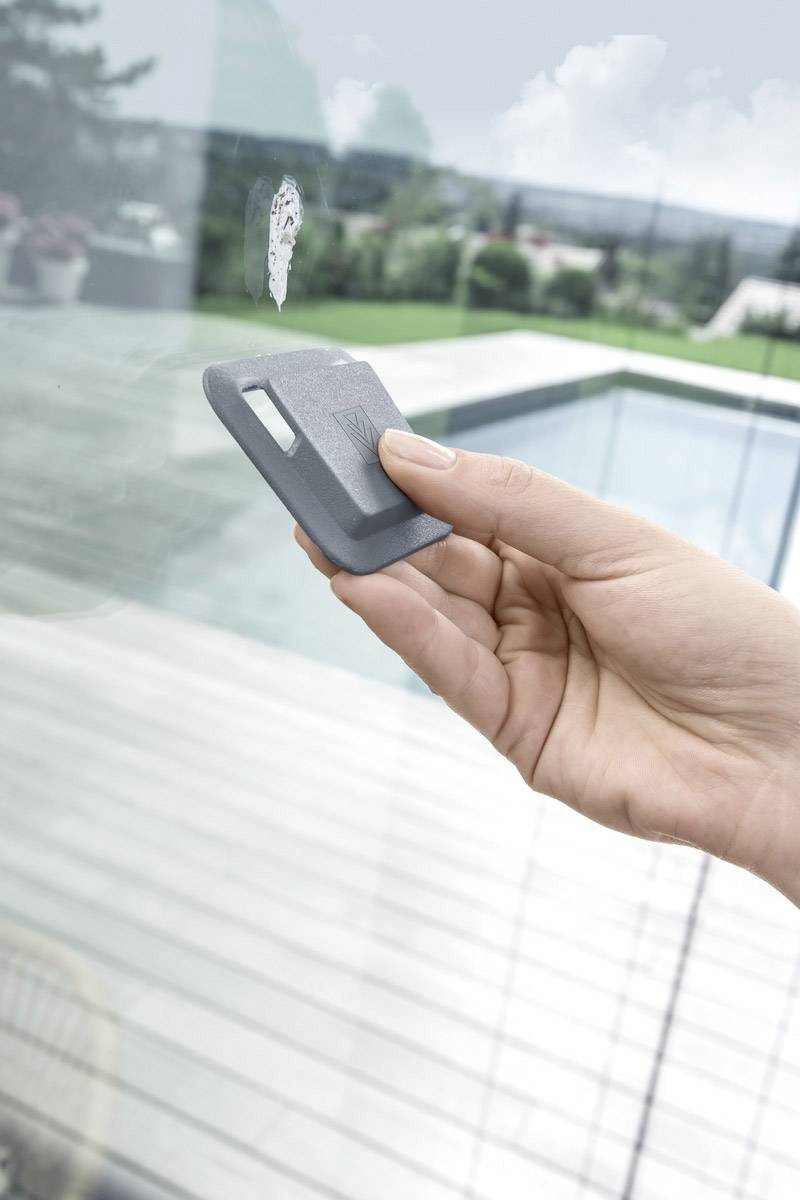 A hand cleans a window pane with a grey cleaning scraper. In the blurred background, a swimming pool and garden landscape can be seen.
