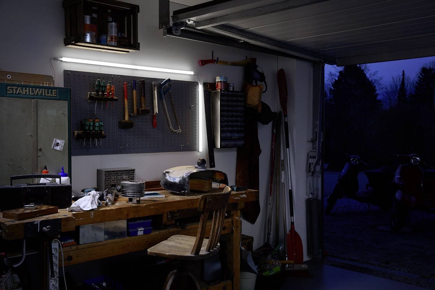 'Workshop at night with open garage door, illuminated workbench, suspended tools and dark exterior view in the background.'