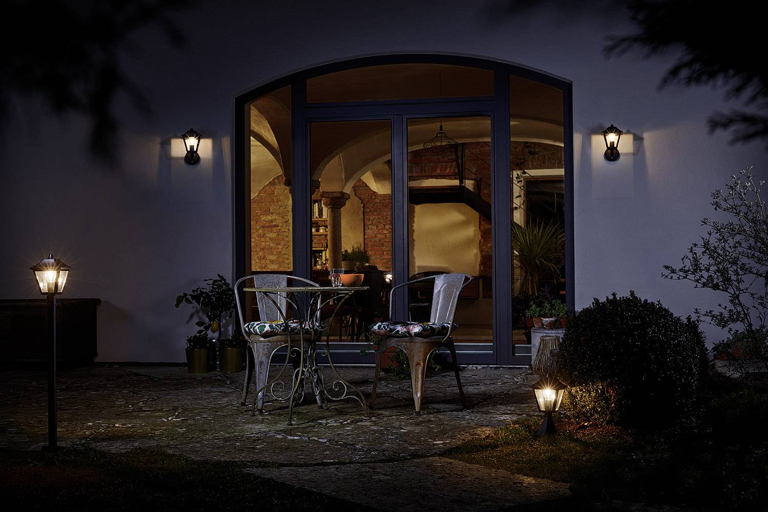 A lit stone garden with two chairs and a round table positioned in front of a large window at night. Lighting emphasises the scene.