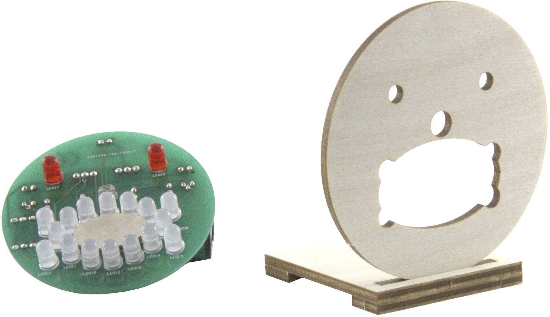 Electronic circuit with LEDs in the shape of a face next to a wooden mounting bracket with a similar facial cut-out.
