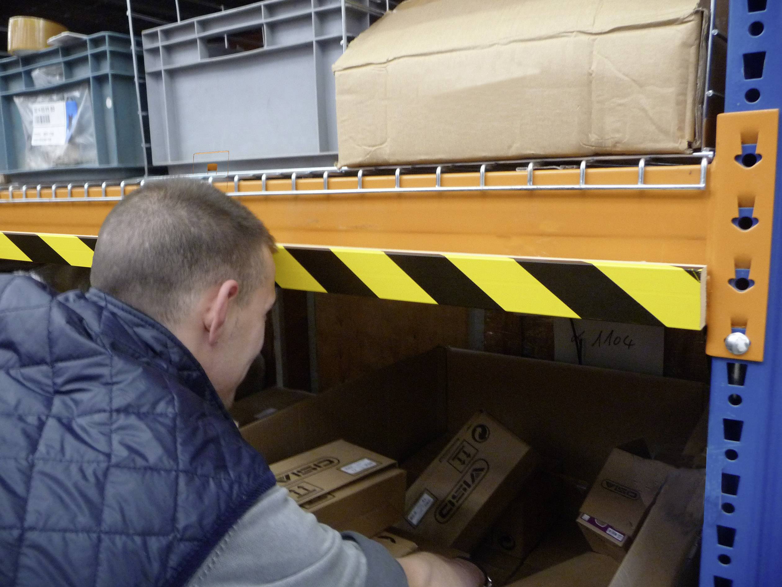 A man is searching for a package on a warehouse shelf. Above him, boxes are stacked on a shelf with yellow and black hazard stripes.