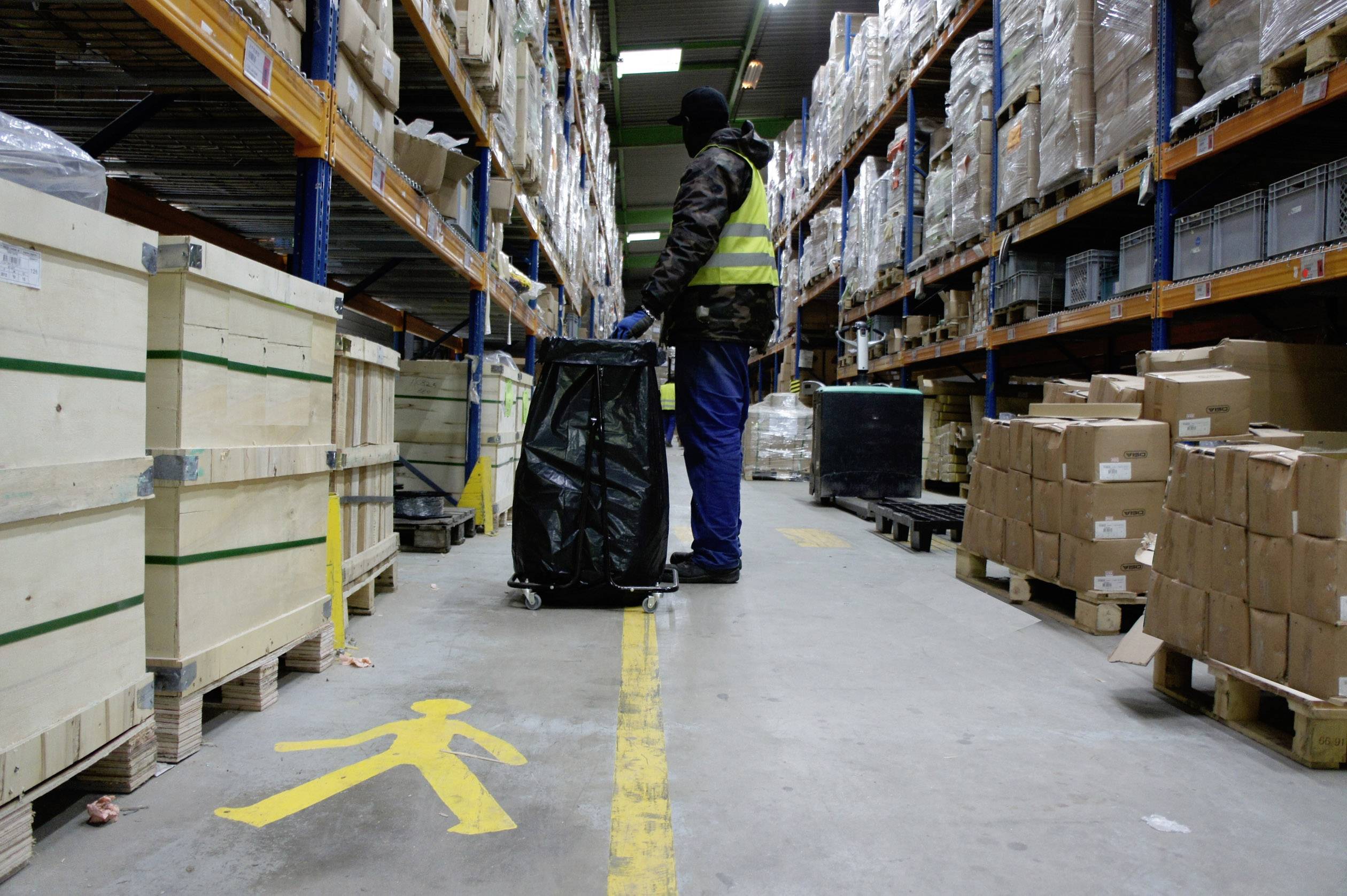 A warehouse worker in a high-visibility vest pulls a loaded trolley through a long warehouse aisle with tall shelves stacked with boxes and crates.