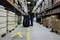 A warehouse worker in a high-visibility vest pulls a loaded trolley through a long warehouse aisle with tall shelves stacked with boxes and crates.