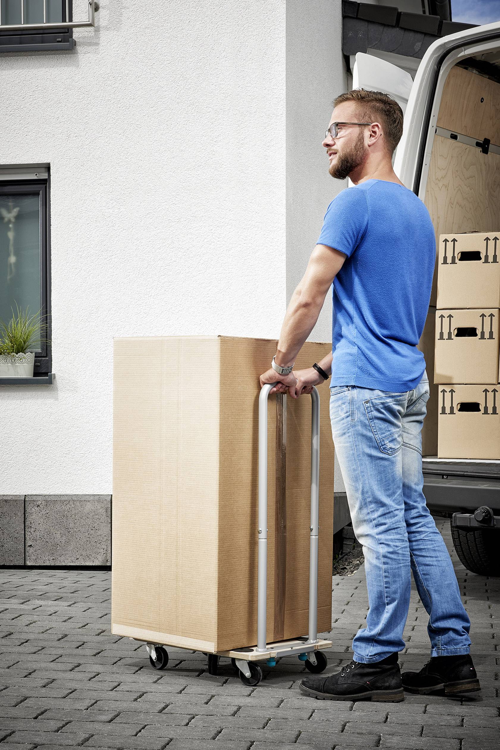 A person in blue clothing is transporting a large cardboard box on a hand trolley. A delivery van is parked in the background.