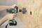 A drill with a green casing is used to bore a round hole in a plywood board.