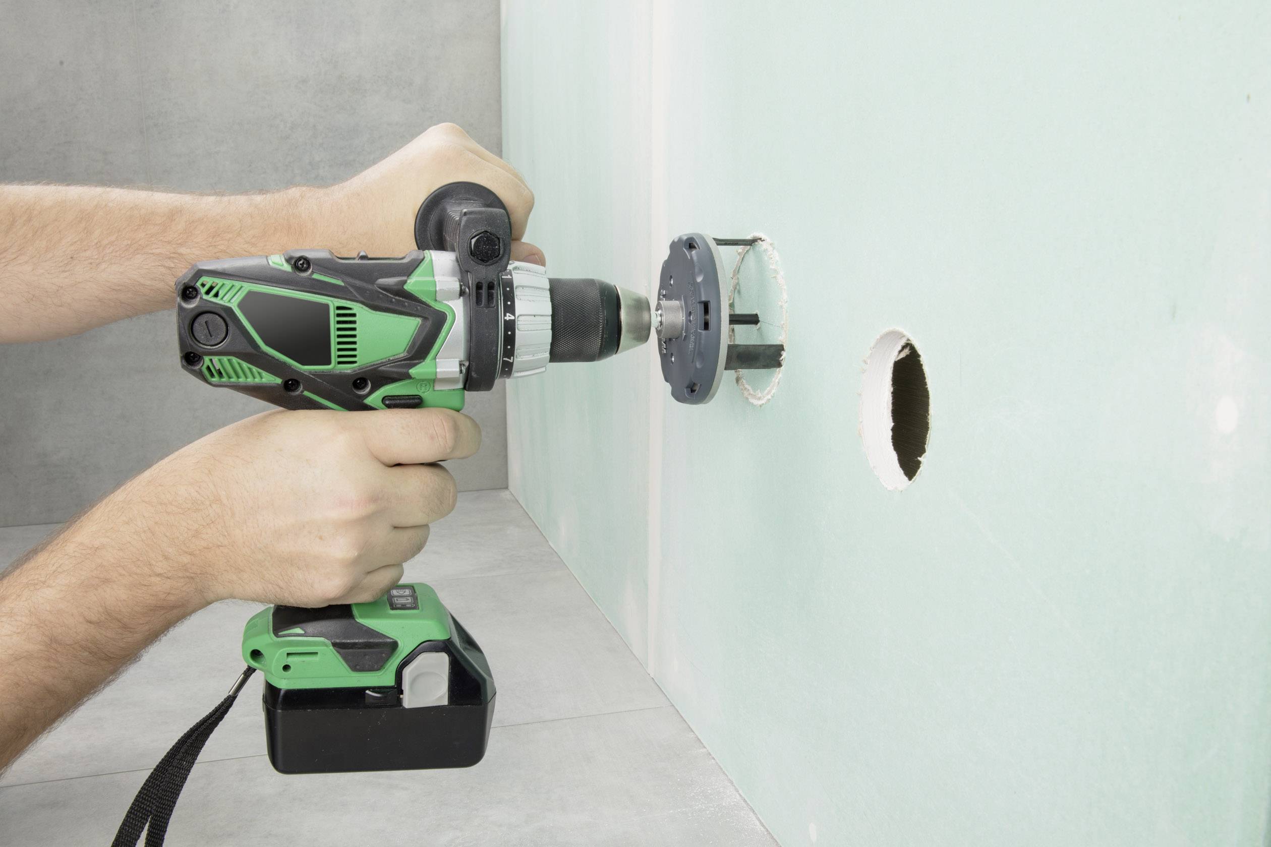 A person is drilling a large round hole in a green wall using an electric drill.