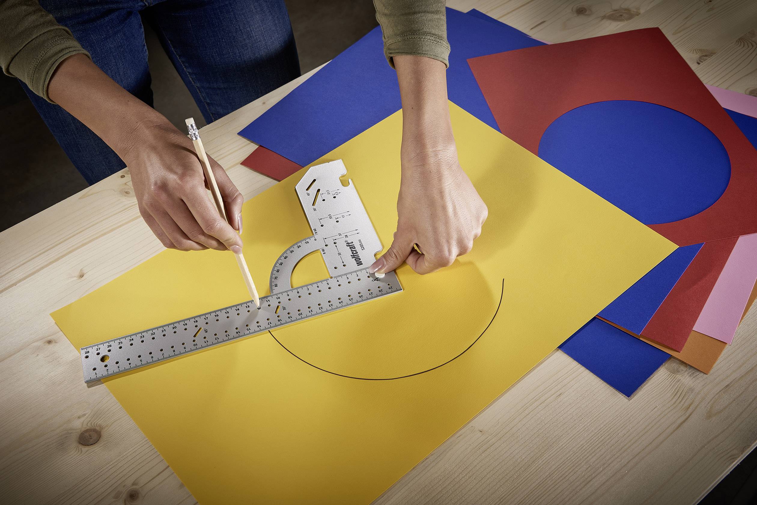 A person is drawing a circle on yellow paper using a ruler and a pencil. Colourful arcs are lying underneath.