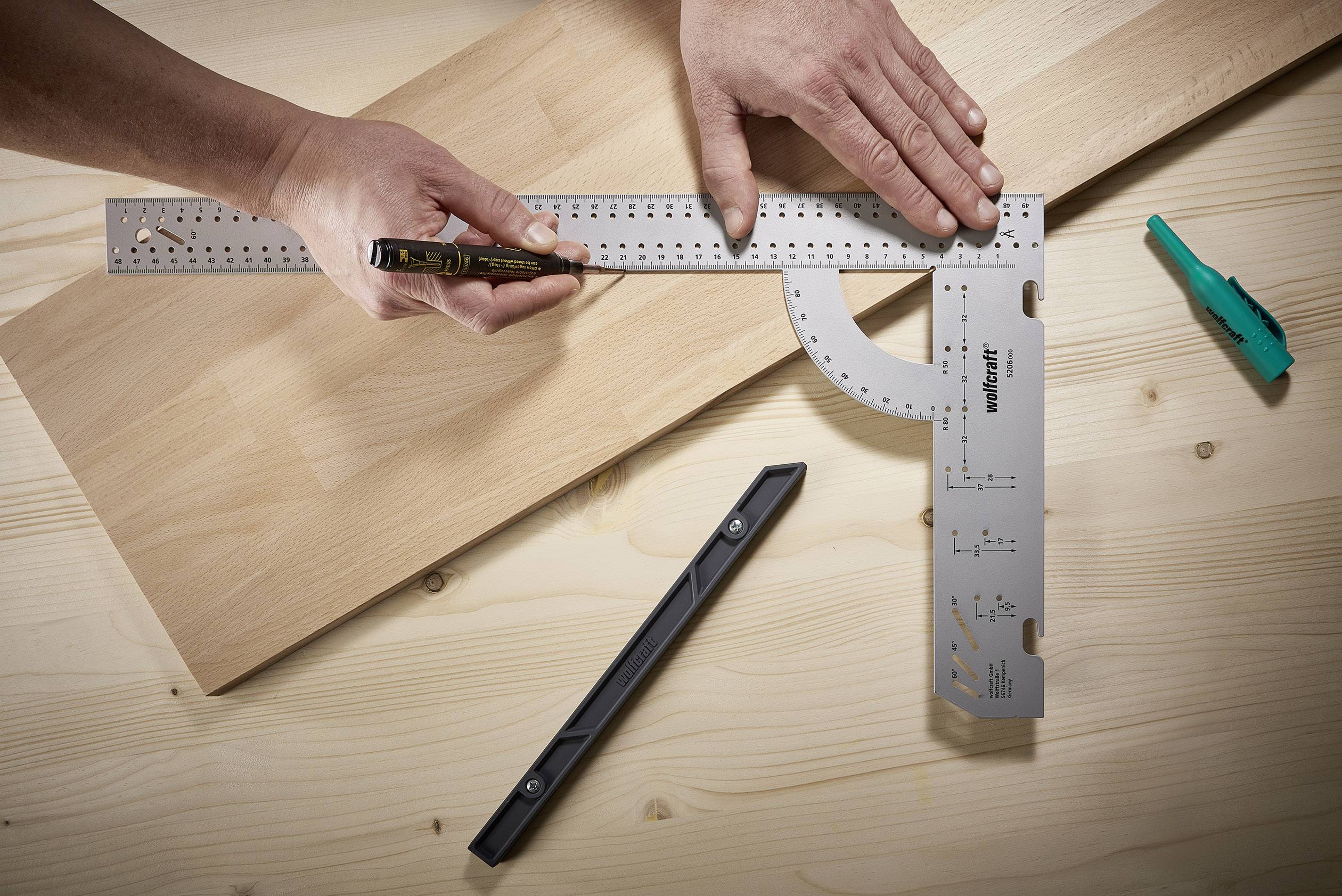 Two hands are using an angle measuring tool and a pencil to mark precise lines on a wooden board. A set square and a knife are lying nearby.