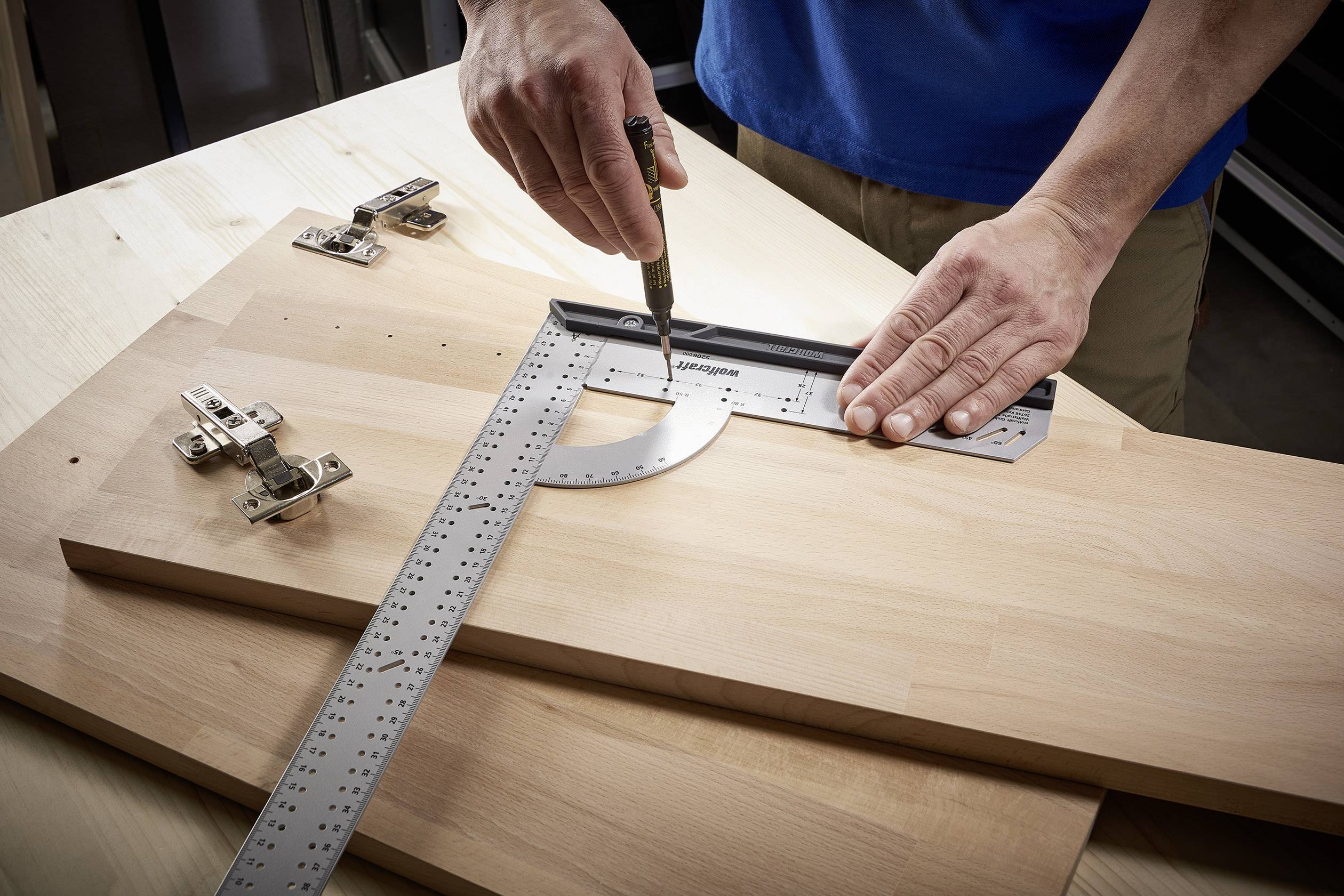 A person is using an angle measuring device to make precise markings on a wooden board. Door hinges are lying next to it on the table.