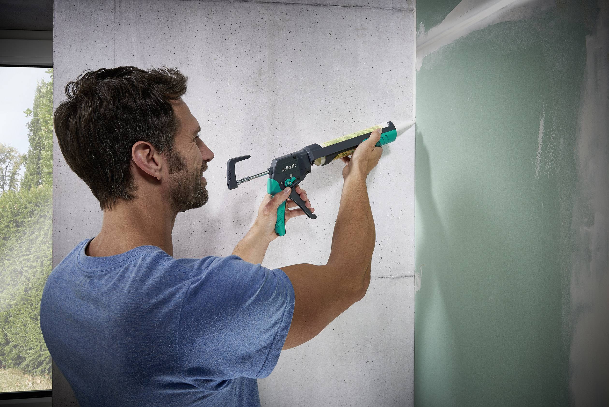 A man is applying sealant to a wall using a cartridge gun. He is wearing a blue T-shirt and working with concentration.
