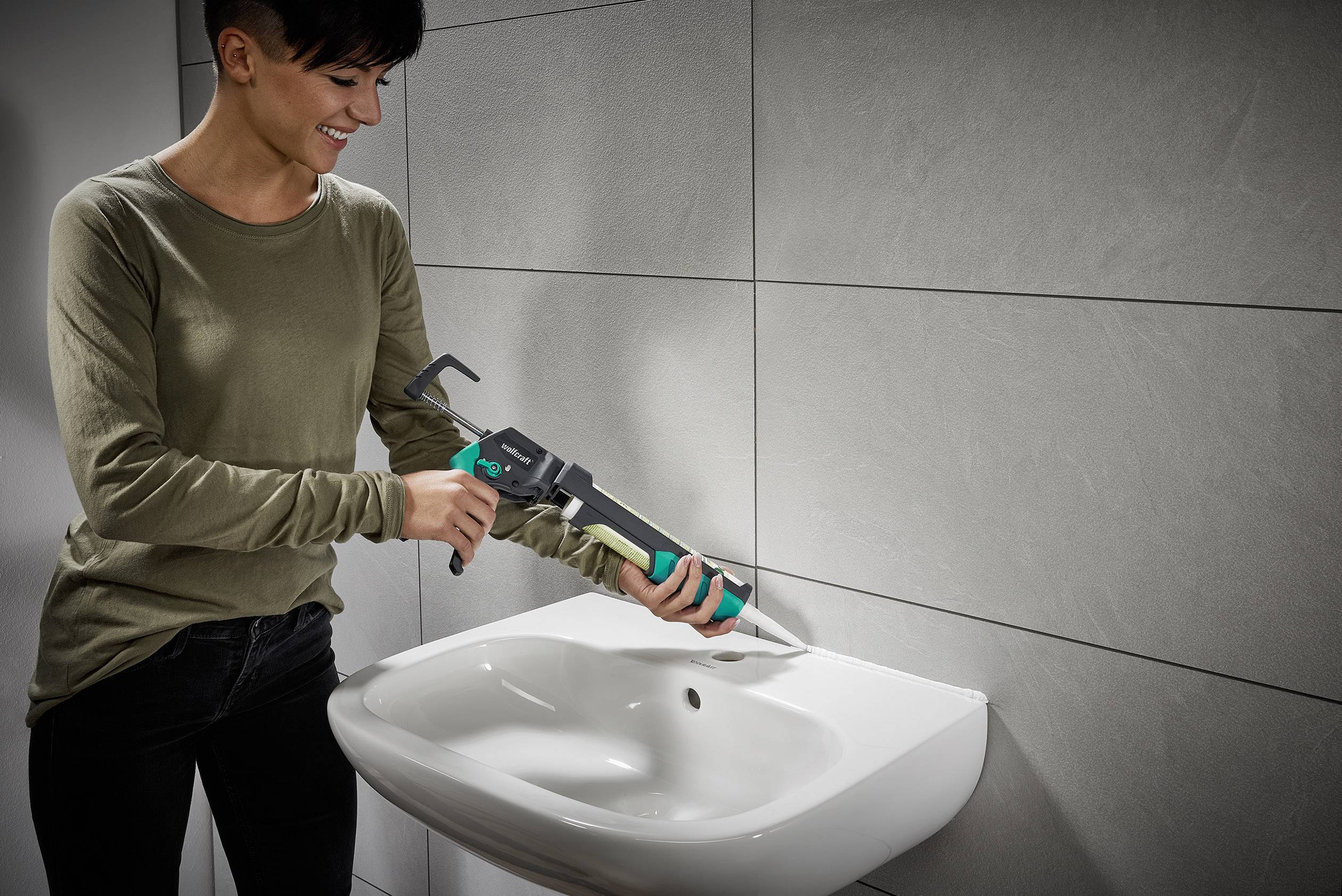 A person is filling the joints around the sink with a cartridge gun. Bathroom tiles are visible in the background.