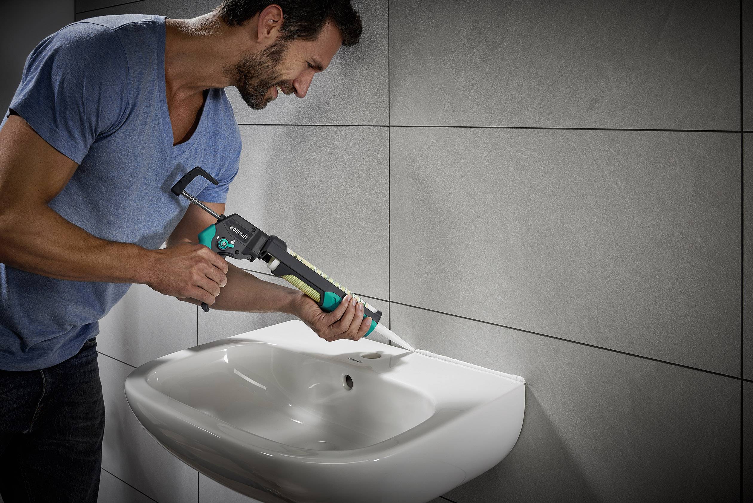A man is applying sealant to the edge of a white sink in a tiled bathroom.