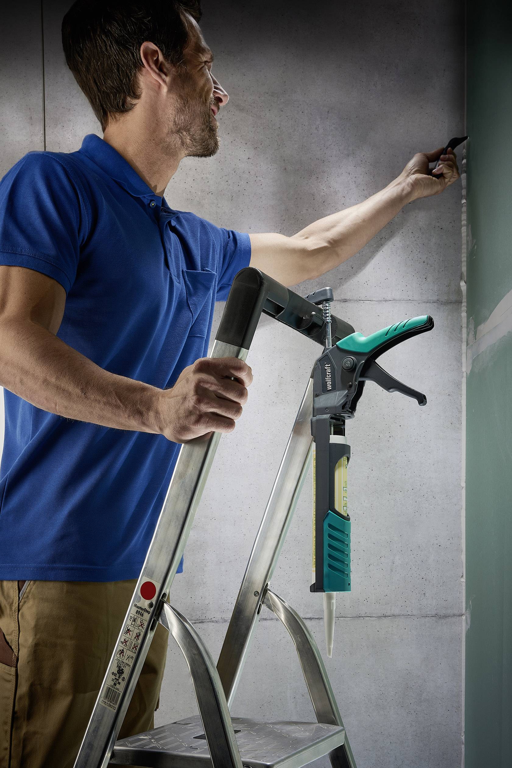 A man is standing on a ladder and painting a wall with a brush. A caulking gun is attached to the ladder.