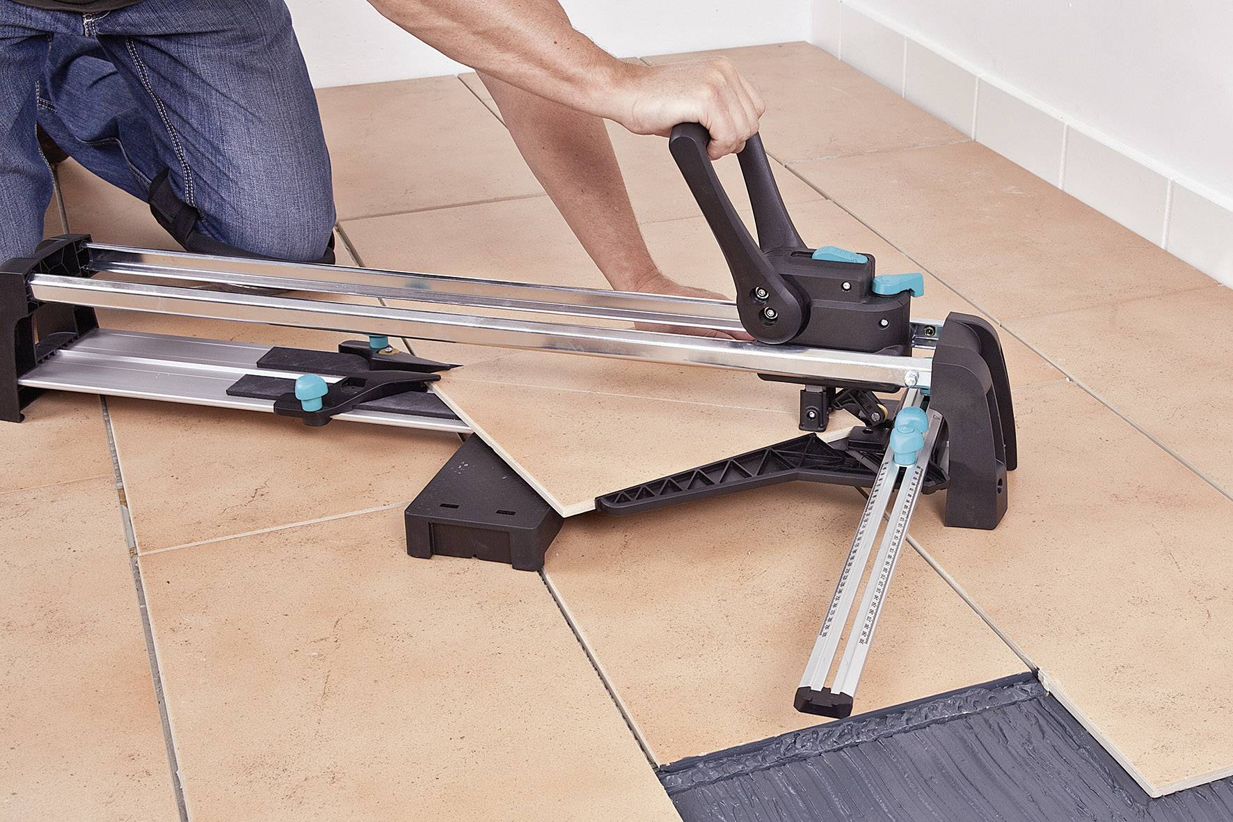 A person is cutting a ceramic floor tile with a manual tile cutter on an already tiled floor.