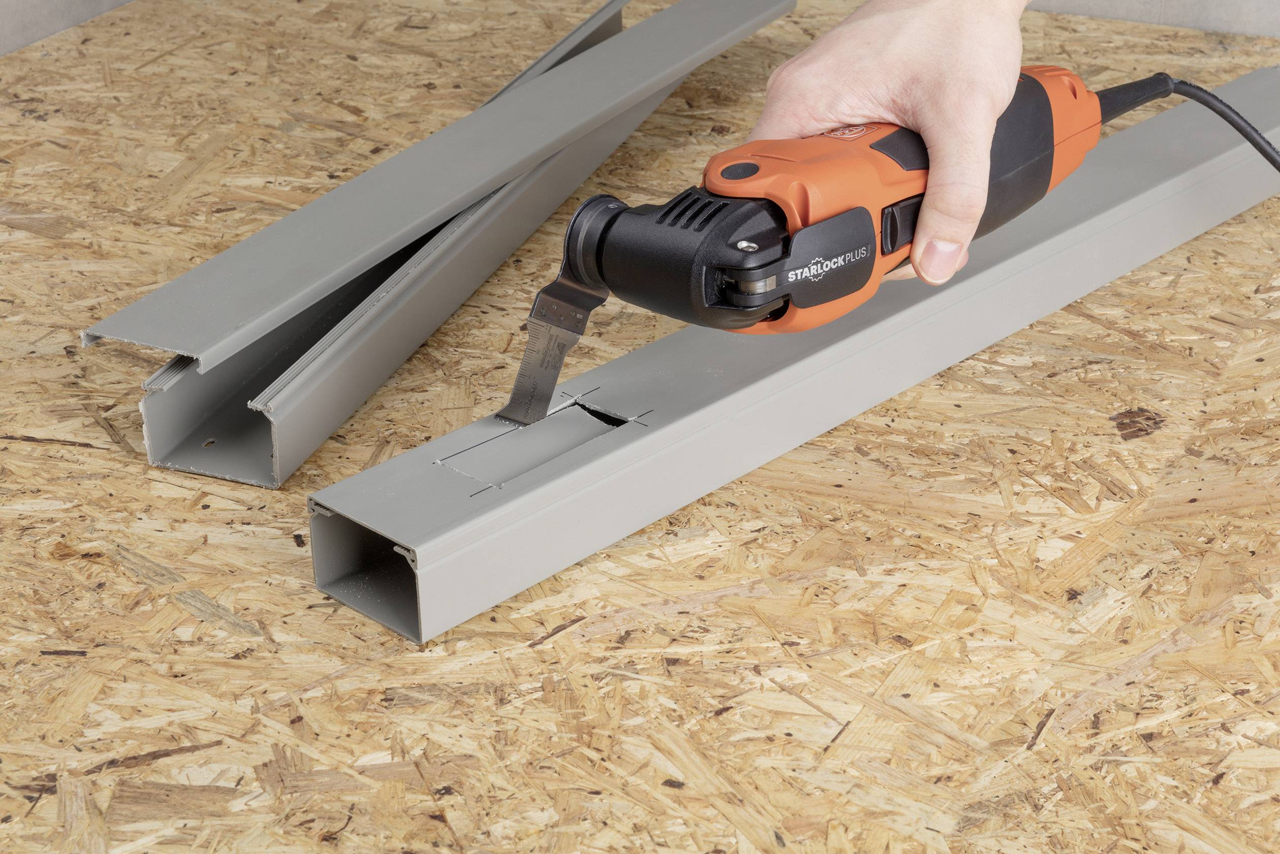 A hand operates an oscillating tool to make a rectangular cut in a grey metal profile.