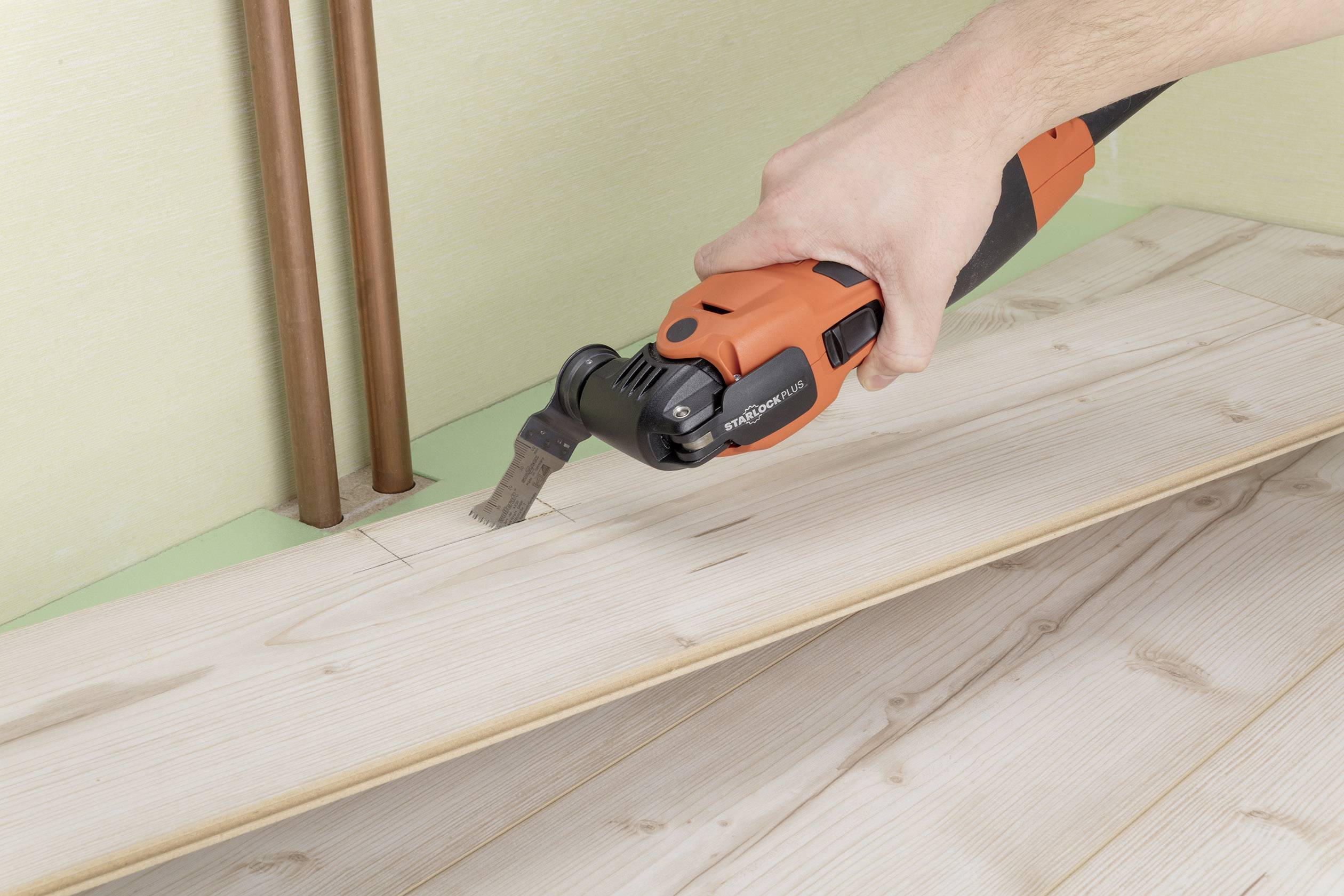 A person is cutting a piece of wood, which is part of floor installation, using a multi-tool. Copper pipes can be seen in the background.
