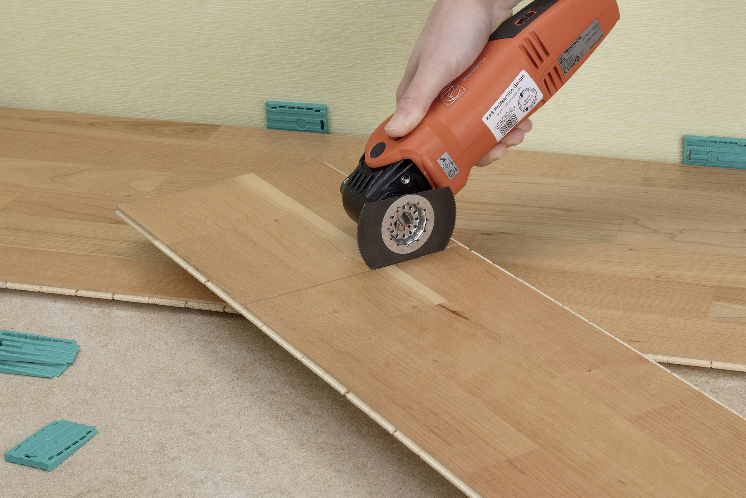 A hand holds an oscillating multi-tool cutting a wooden board. Several spacers are lying on the ground.