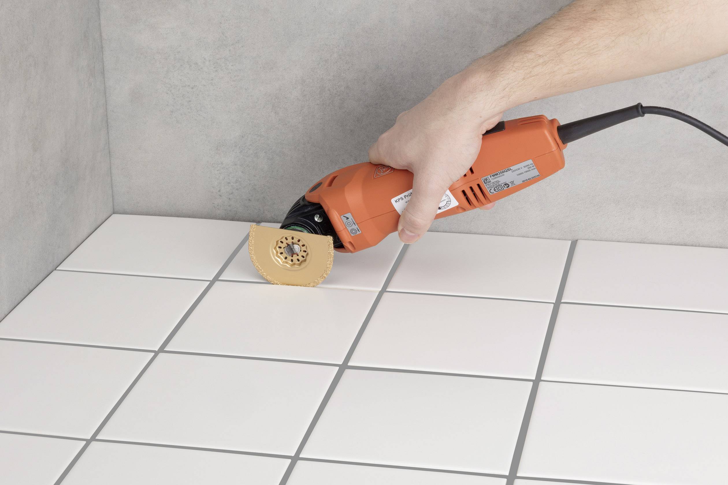 A hand is holding an oscillating multi-tool to work on the joints between white tiles on a floor surface.