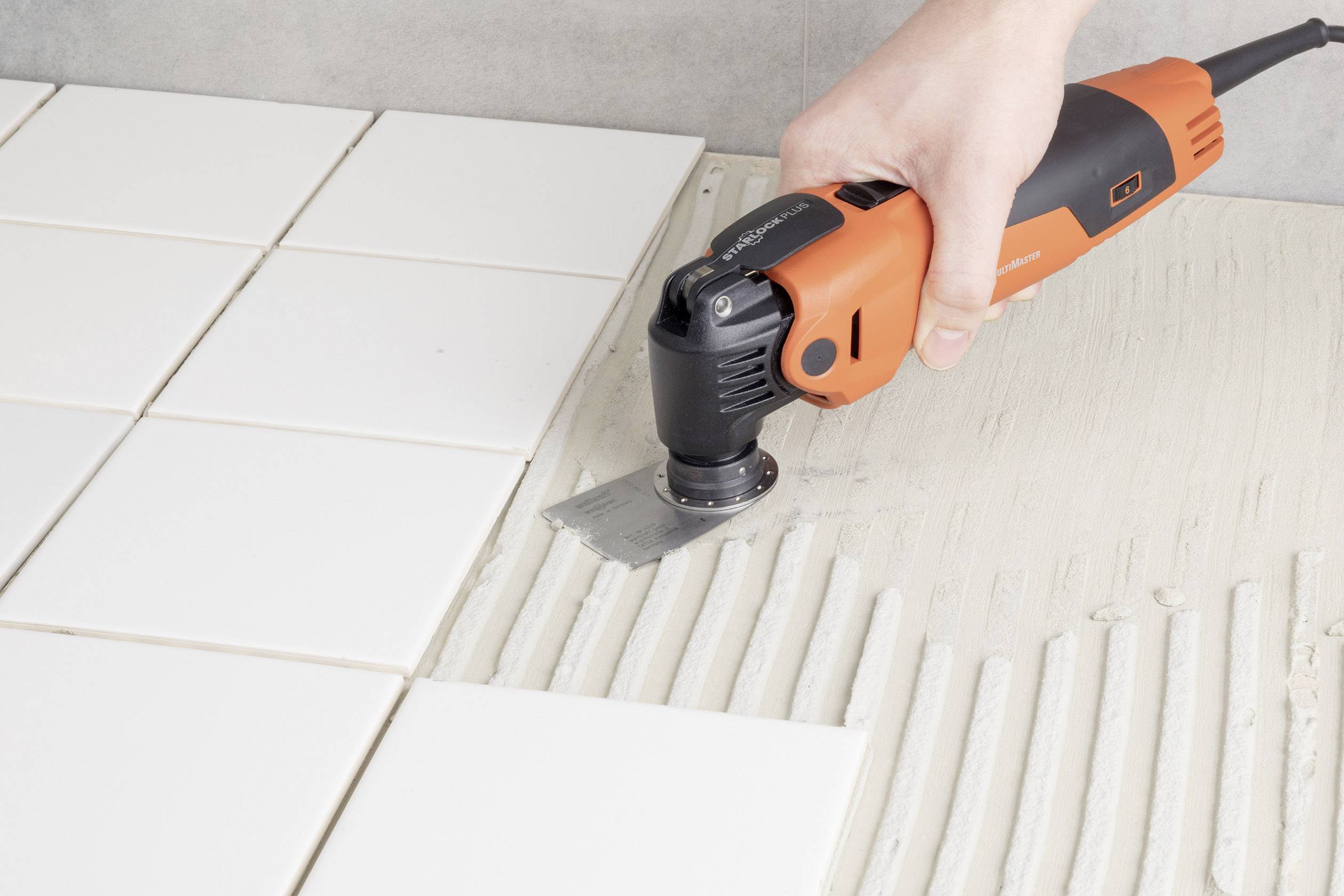 A hand is holding an orange power tool removing adhesive between white tiles.
